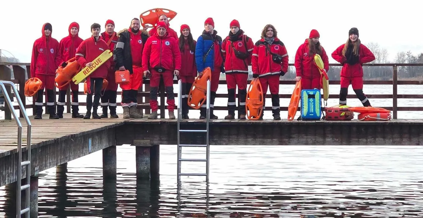 Kilkanaście osób z Młodzieżowej Grupy WOPR w czerwonych kurtkach stoi na pomoście nad jeziorem, ze sprzętem ratowniczym: bojami SP, deską z napisem Rescue, defibrylatorem AED i torbą z tlenem